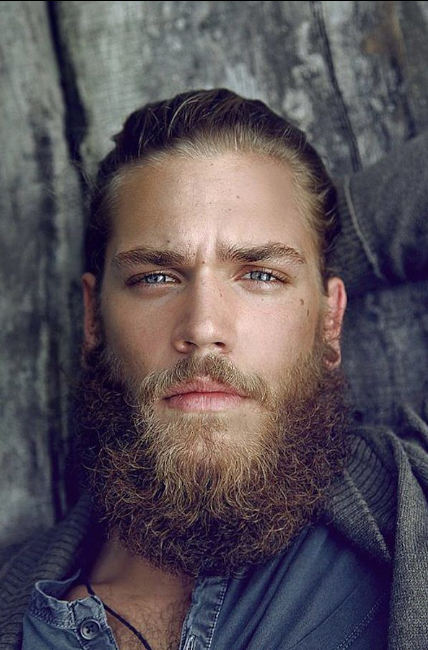 Man with a beard and tattoos on his neck against a textured wall.